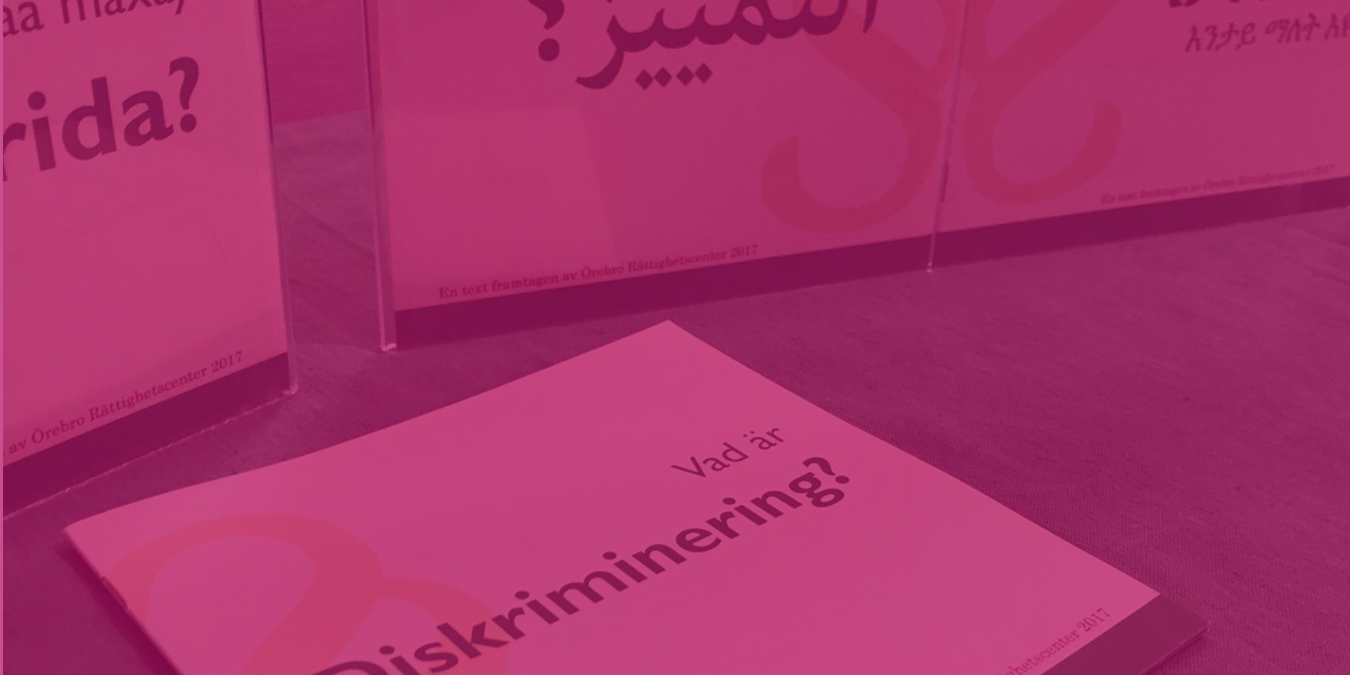 Vad är diskriminering rosa En broschyr med rubriken Vad är diskriminering? I bakgrunden syns fler broschyrer med samma rubrik fast på somaliska och arabiska.