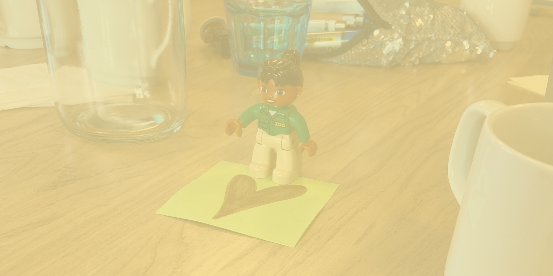 kvinnodagen debatt webb En kvinna av lego med uppsatt hår och en zoo-skötartröja som står på en post-it med ett ritat hjärta på ett bord med kaffekoppar.